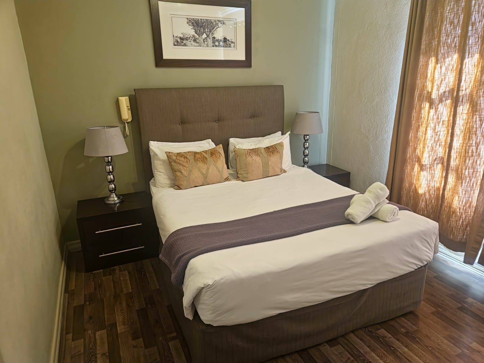 Habitación de hotel con cama, dos mesitas de noche, lámpara, un cuadro en la pared y ventana con cortinas.