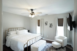 Quarto com cama, uma cadeira, ventilador de teto e duas janelas com cortinas.