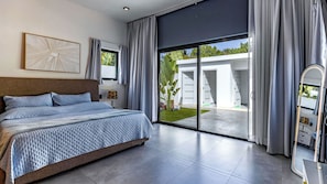 Ein modernes Schlafzimmer mit einem großen Bett, einem Nachttisch, einer Lampe und Blick auf ein Haus durch eine Schiebetür.