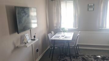 A room with a flat-screen TV mounted on the wall, a dining table with chairs, a fluffy rug, and a window with curtains.