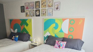 Ein Doppelbett mit weißer Bettwäsche und einem farbenfrohen Kopfteil, ein Nachttisch mit Lampe sowie eine Wand mit gerahmten Illustrationen aus Kinderbüchern.