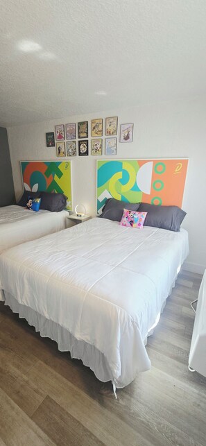 A double bed with white bedding and a colorful headboard, a bedside table with a lamp, and a wall with framed children's book illustrations. - Cozy 2-Queen Room, Pool, Near Disney & Attractions. (Kissimmee)