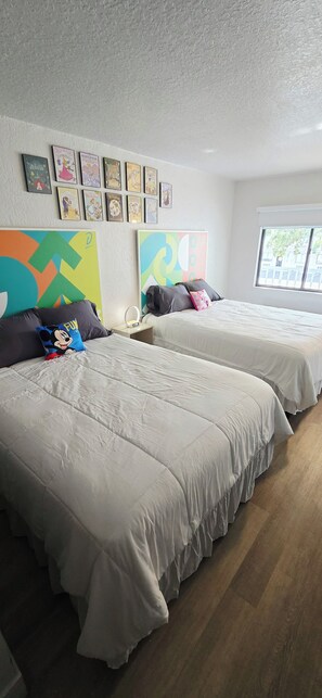 A hotel room with two beds, a colorful abstract painting, and a window with a view. - Cozy 2-Queen Room, Pool, Near Disney & Attractions. (Kissimmee)