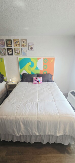 A bedroom with a bed, pillows, a nightstand, a wall clock, and a series of framed posters. - Cozy 2-Queen Room, Pool, Near Disney & Attractions. (Kissimmee)