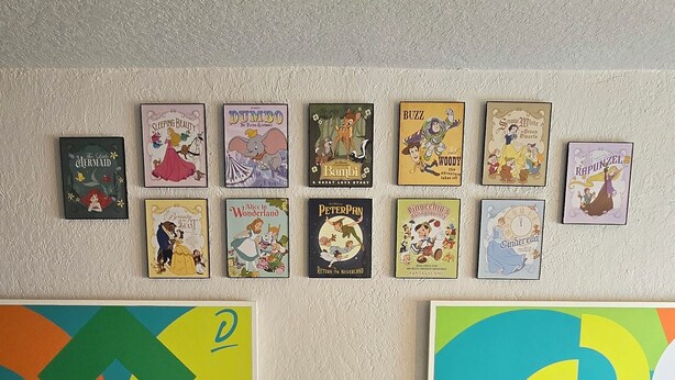Ein Zimmer mit zwei Betten, einem Nachttisch mit einer Uhr und einer Wand, die mit Illustrationen aus Kinderbüchern verziert ist.