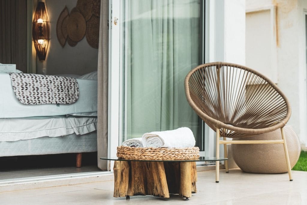 Une pièce comprenant un lit, une chaise en osier, un panier rond en osier et une table en verre sur laquelle est posée une serviette enroulée.