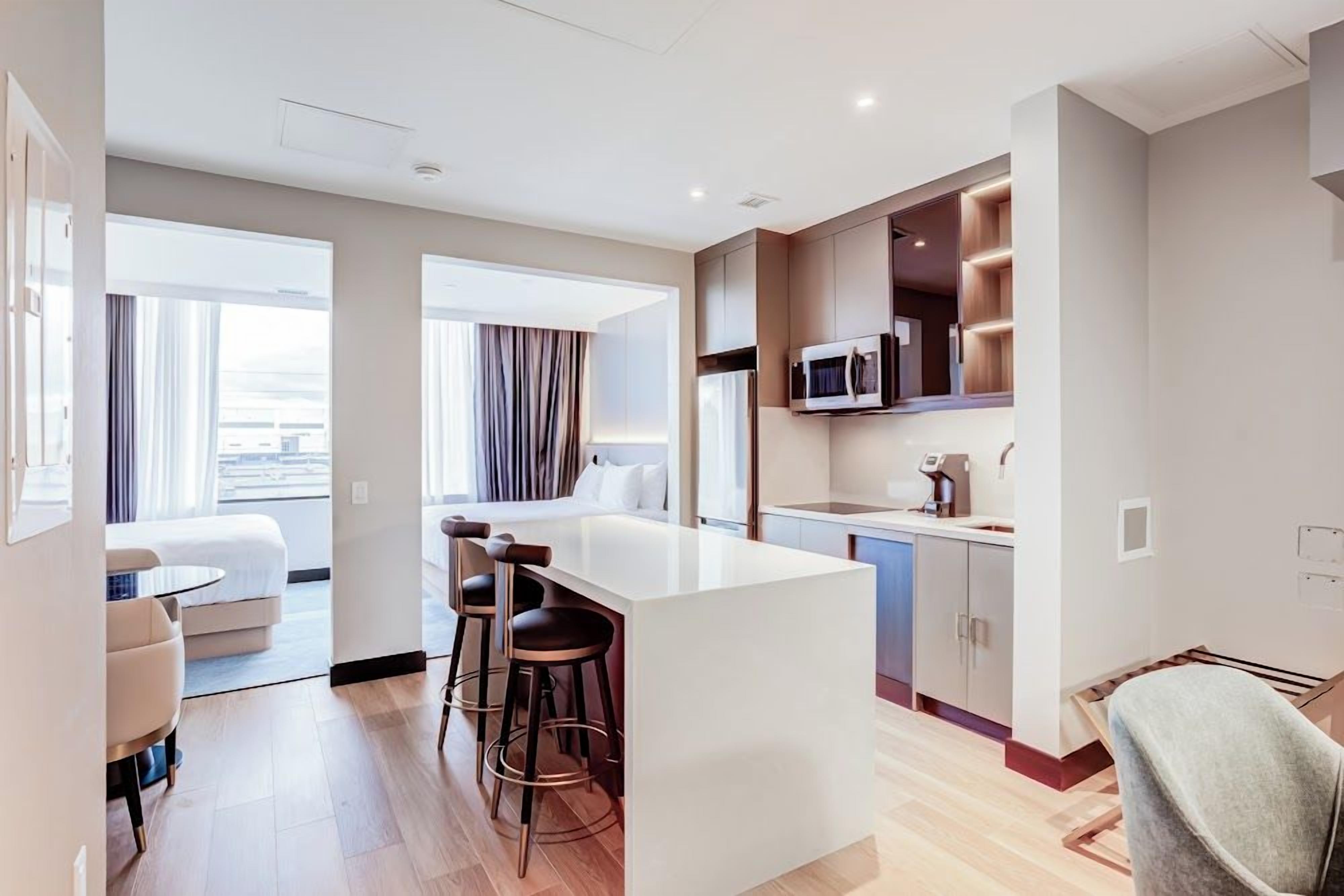Una habitación de hotel moderna con kitchenette, una zona de comedor con taburetes de bar, un ventanal con cortinas y una sala de estar separada con sofá y mesa de centro.