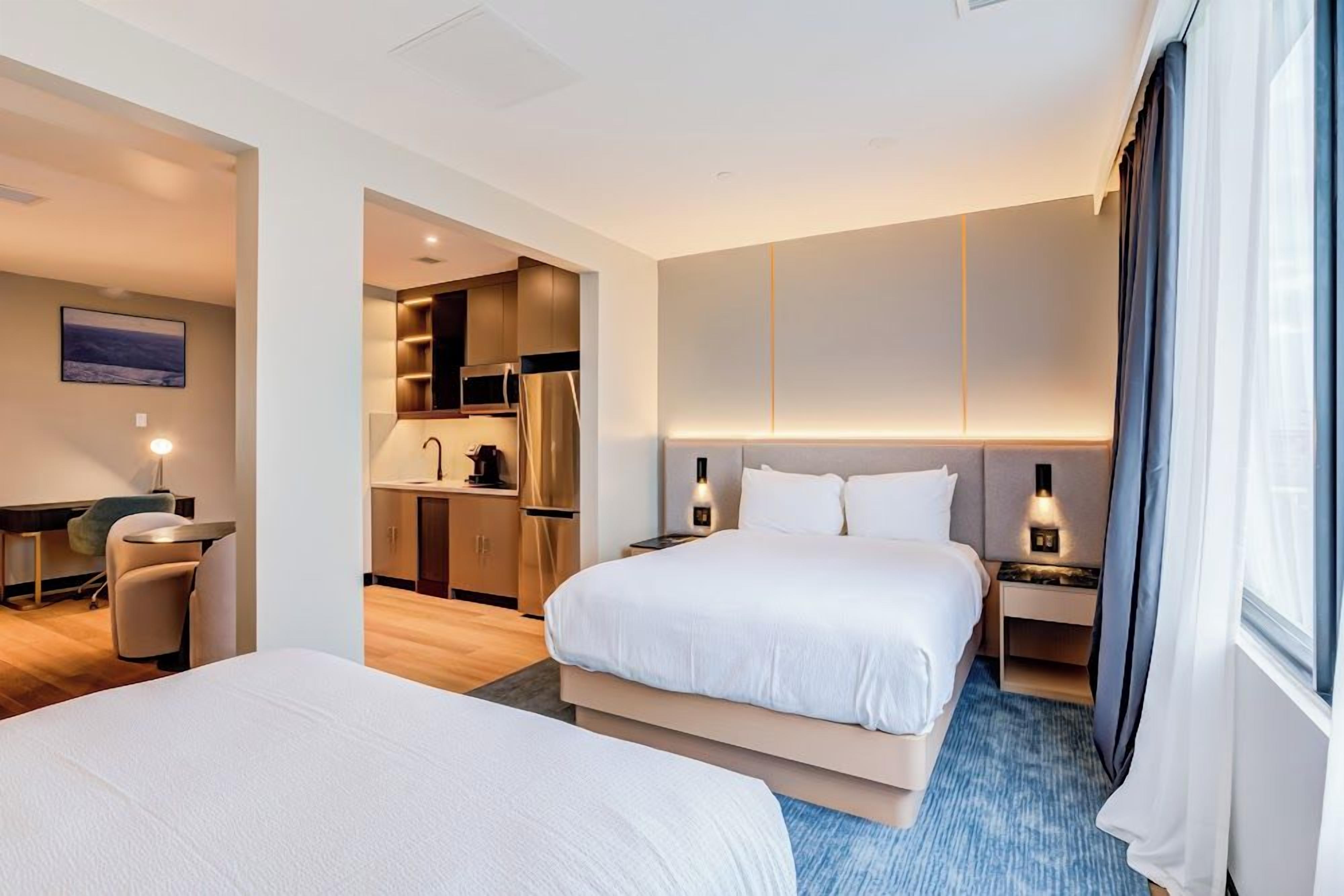 A hotel room with a bed, bedside tables, a desk, and a kitchenette.