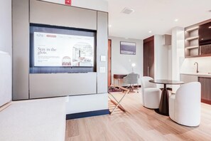 Una habitación de hotel moderna con una pantalla grande mostrando un anuncio, una zona de comedor con una mesa redonda y sillas, y una cocina con armarios y un lavabo.