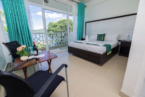 A hotel room with a balcony, a bed with white bedding, a wooden table with a chair, and a potted plant.