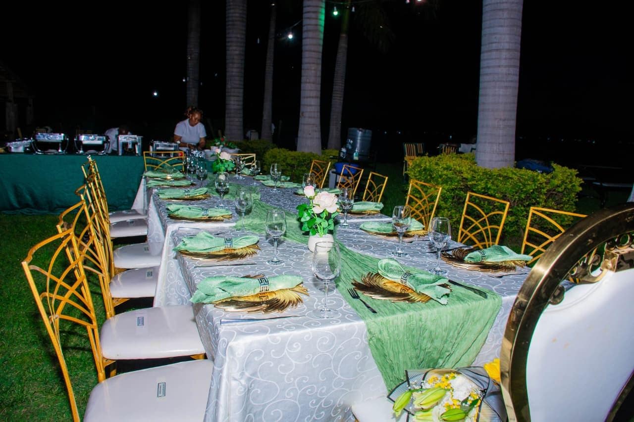 Mesa de comedor al aire libre con sillas dispuestas para varios comensales y una persona en el fondo.