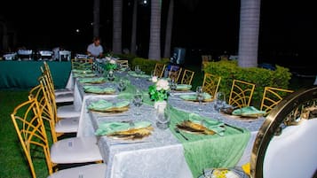 Mesa de comedor al aire libre con sillas dispuestas para varios comensales y una persona en el fondo.