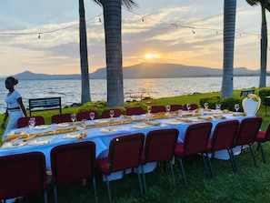 An outdoor dining setup with a table set for multiple guests, chairs, and a view of the sunset over a body of water.