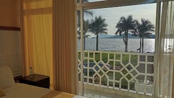 Habitación de hotel con una ventana grande, cama, mesita de noche y vista al mar y palmeras.