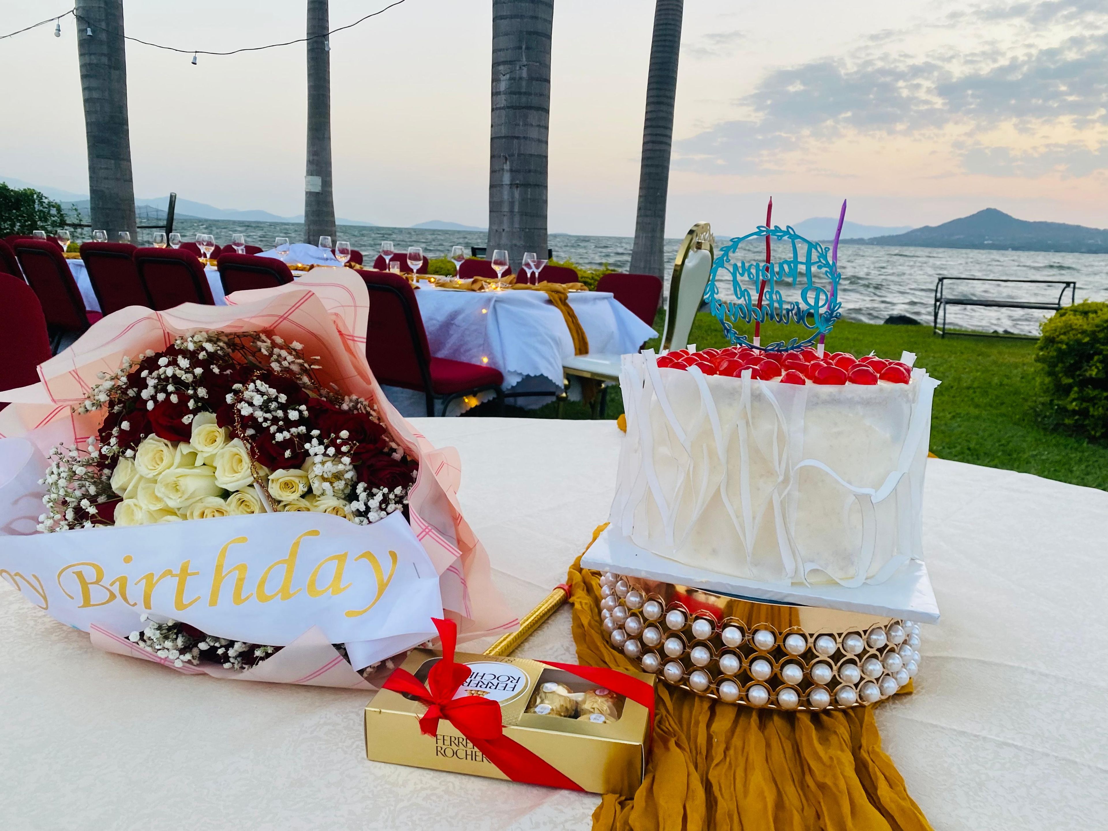Une table ornée d’un bouquet d’anniversaire et d’un gâteau décoré, le tout mis en valeur par un décor de palmiers et d’une étendue d’eau.