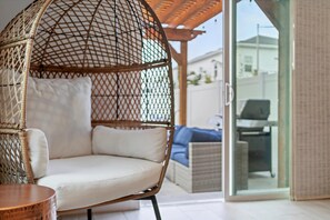 Ein Korbstuhl mit Kissen, ein Holztisch und Blick auf ein blaues Sofa sowie ein weißes Gebäude durch eine Glastür.