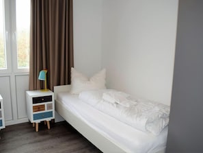 A single-bed room with a white bed, a nightstand with drawers, a lamp, and a window with curtains. - House No. 25, Holiday Home Fischerhus (Wangerland)