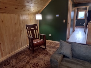 A room with a wooden ceiling, a green wall, a wooden chair, a patterned rug, and a grey sofa.