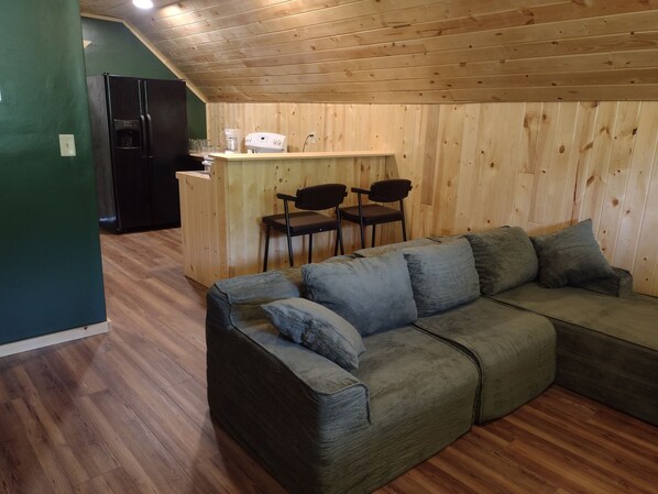 A cozy room with a wooden ceiling, a green wall, a sofa with cushions, a kitchen area with a refrigerator and a coffee maker, and a bar with chairs. - Welcoming 1-bedroom apartment in charming Squaw Lake (Squaw Lake)