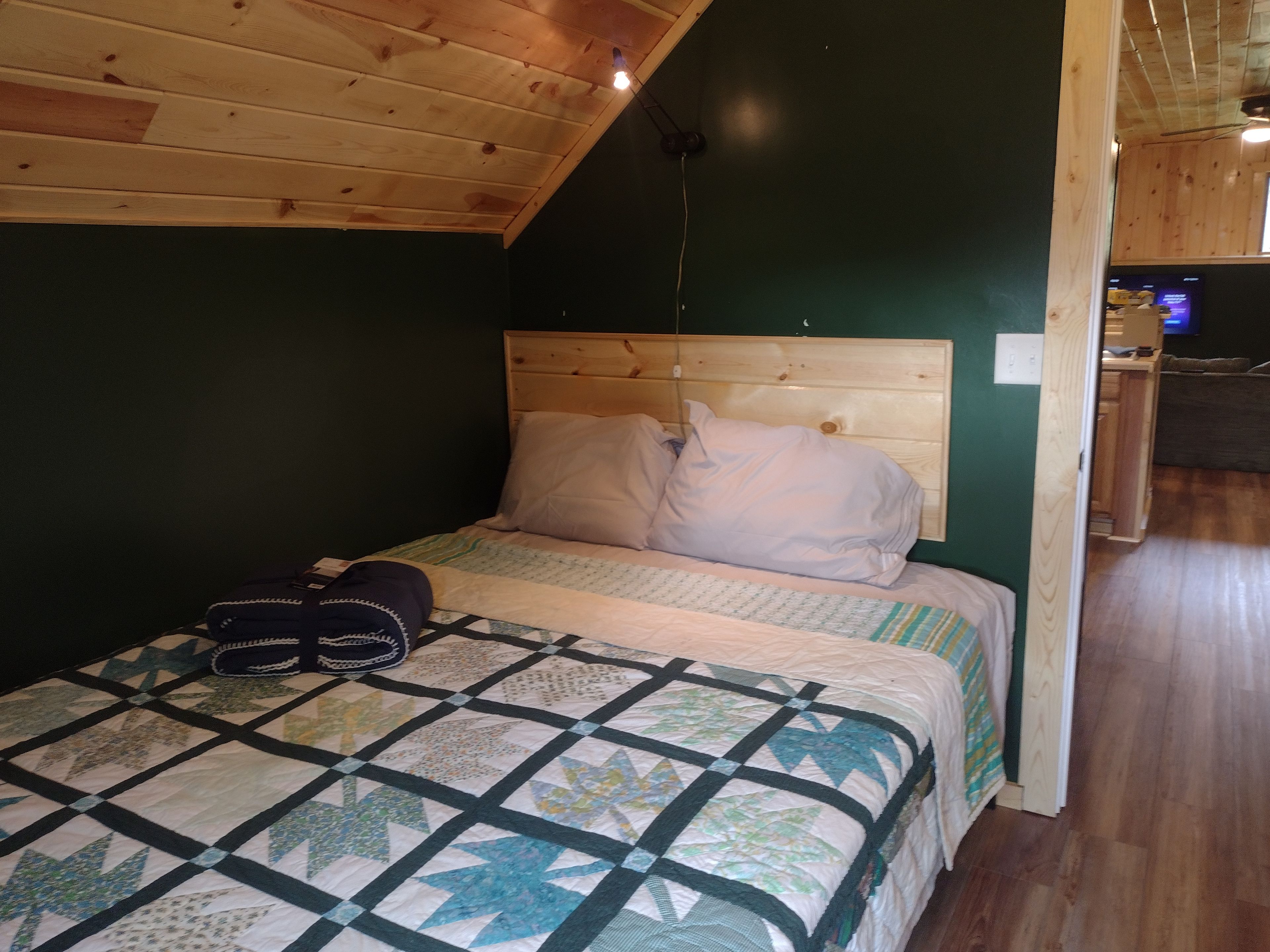 A bedroom with a wooden ceiling, a bed with a quilt, pillows, and a pair of shoes on the floor.