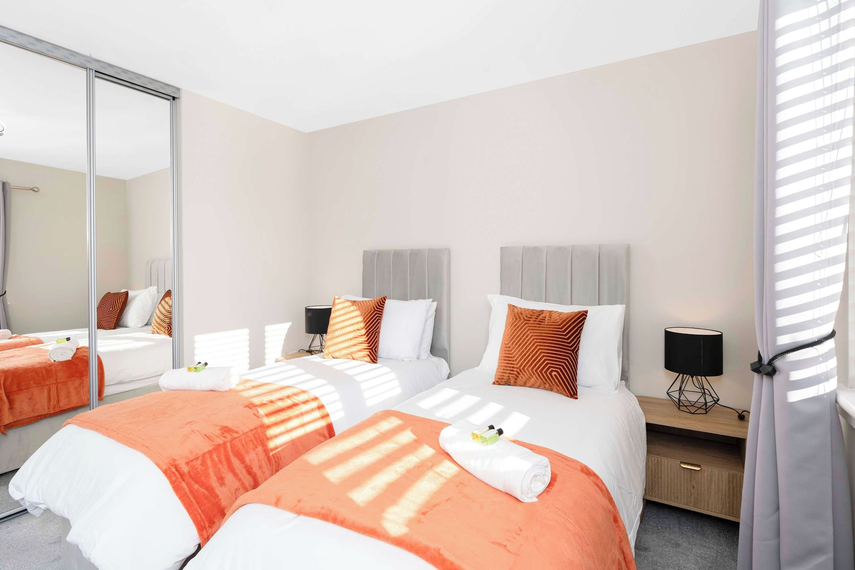 A hotel room with two beds, a large mirror, a bedside table with a lamp, and a window with blinds.