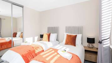 A hotel room with two beds, a large mirror, a bedside table with a lamp, and a window with blinds.