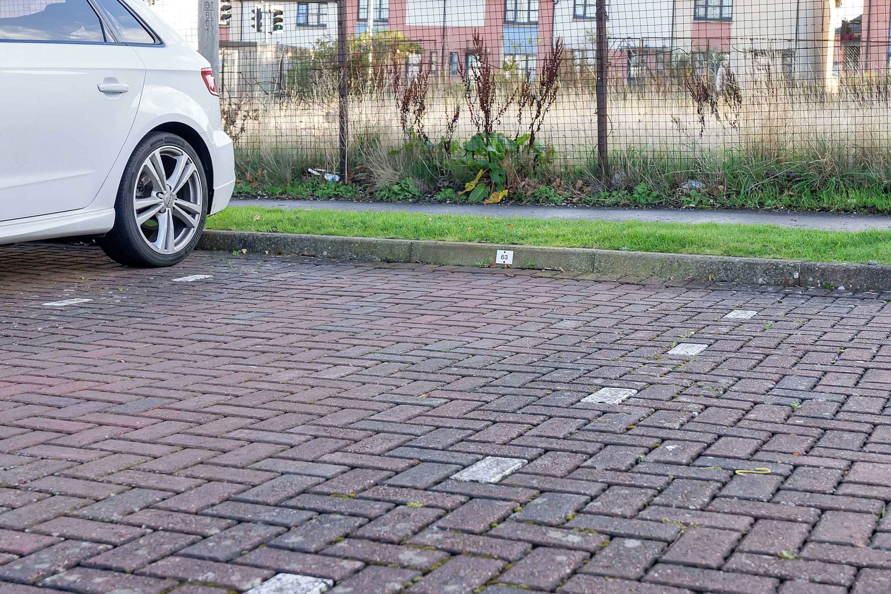 En vit bil parkerad på en asfalterad yta, intill en trottoarkant, med en liten skylt med siffran 63.