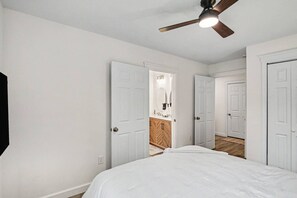 Une chambre à coucher avec un lit, un ventilateur de plafond et une salle de bain visible à travers une porte ouverte.