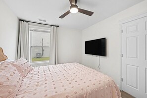 Un dormitori amb un llit, un ventilador de sostre, una finestra amb cortines i un televisor penjat.