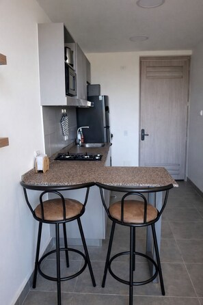 Una cocina compacta con encimera de granito, dos taburetes de bar, horno empotrado y refrigerador.