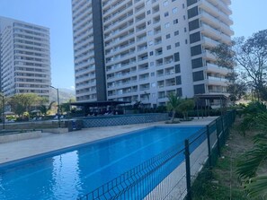 Ein Swimmingpool mit umgebender Sicherheitszäunung, neben einem Hochhaus mit mehreren Balkonen.