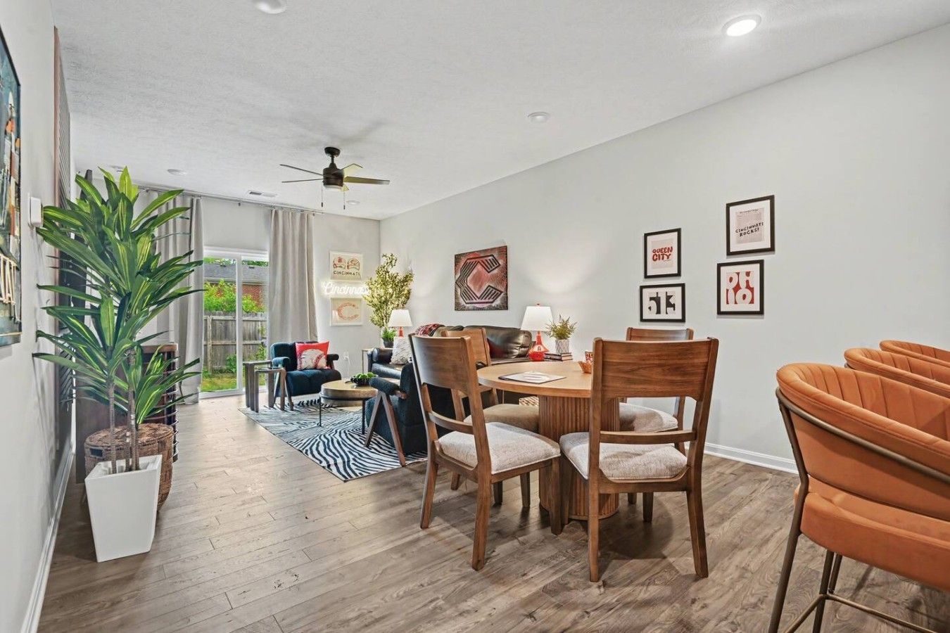 A modern living room with a dining area, featuring a ceiling fan, a large potted plant, and a view into a balcony.