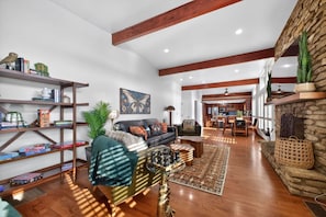 A spacious living room with a fireplace, wooden beams, and a kitchen in the background.