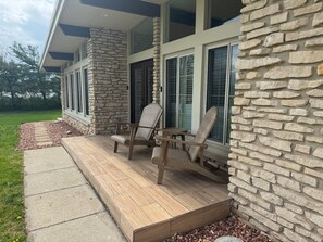 A covered porch with two wooden chairs and a small table, stone walls, and a window with blinds.