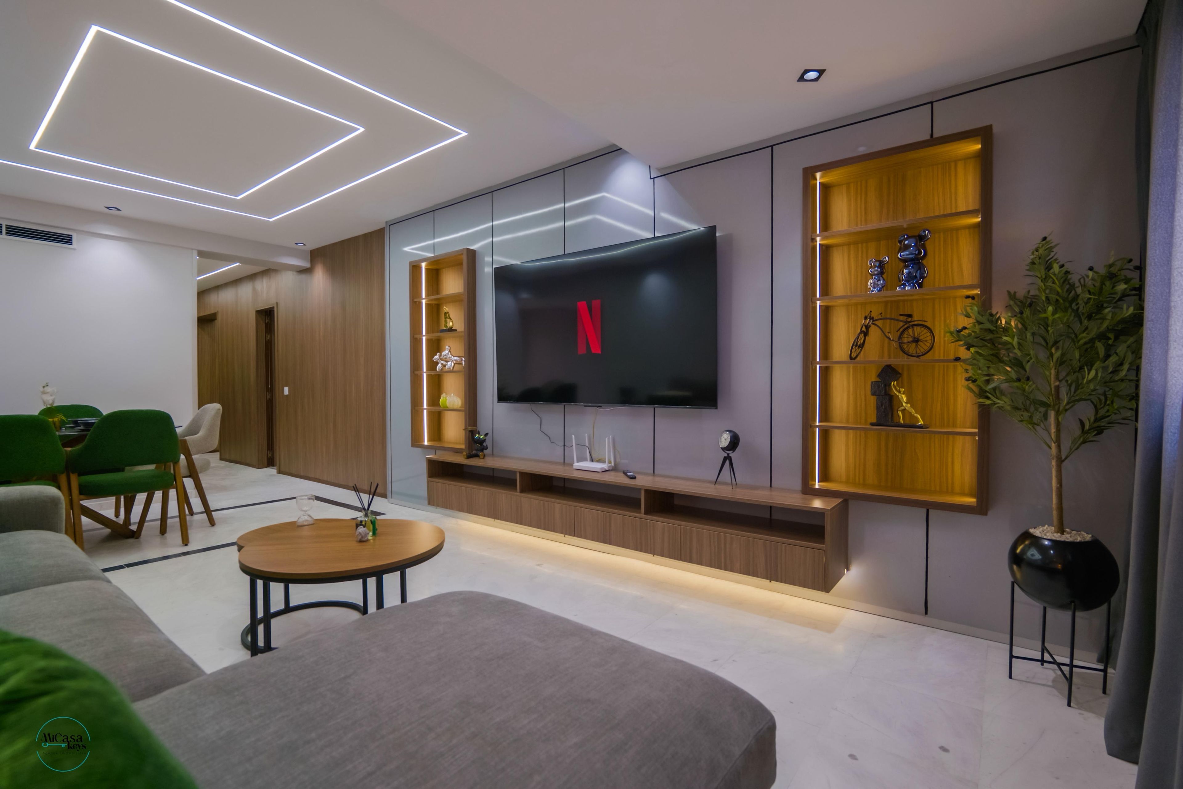 A modern living room with a flat-screen TV mounted on the wall, wooden shelving with decorative items, a green sofa, and a small round coffee table.