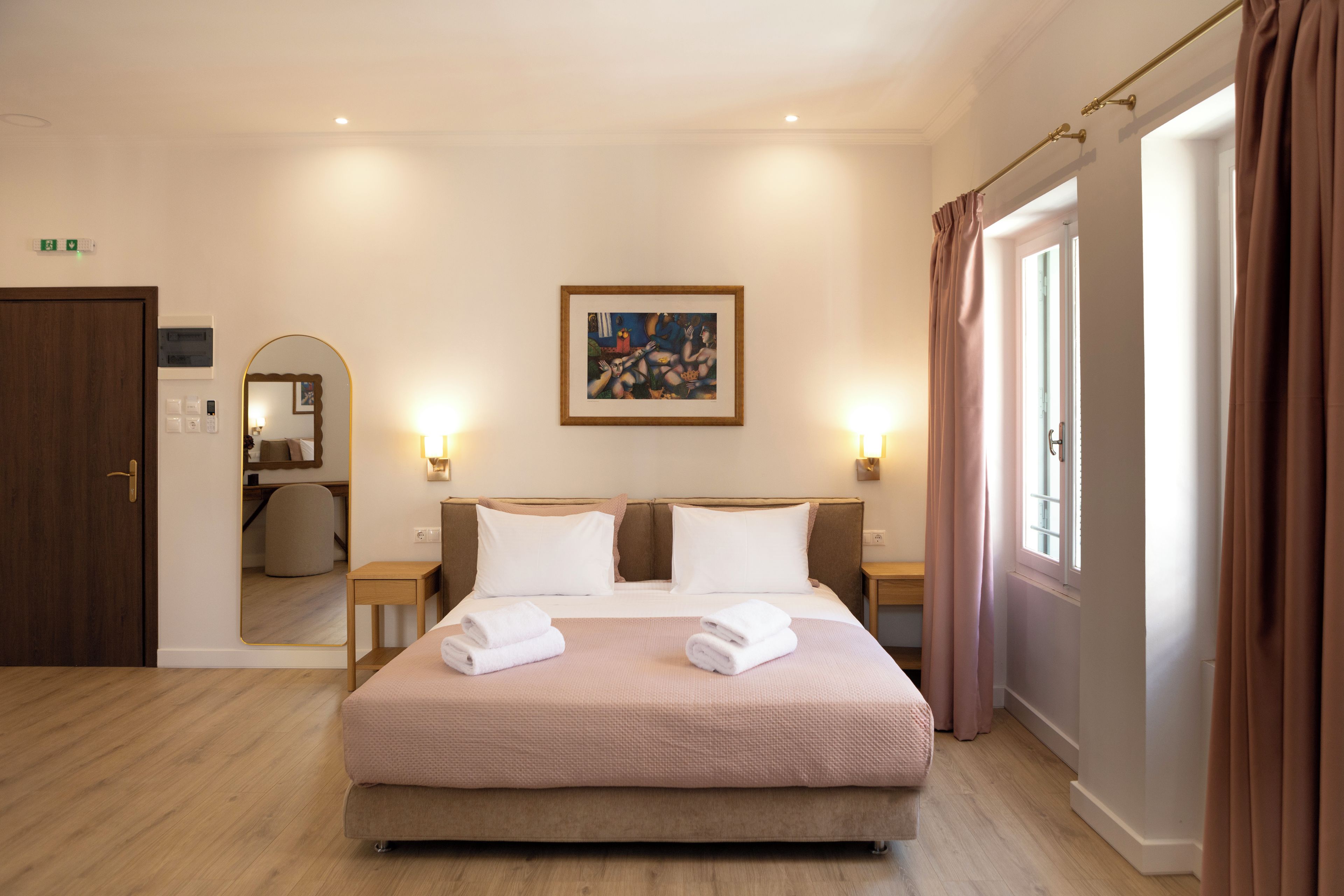 Habitación de hotel con una cama, dos mesitas de noche, un cuadro en la pared y una puerta con letrero de salida de emergencia.