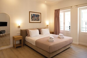 A hotel room with a bed, bedside tables, a painting, a television, and a window with curtains. - Aioliki Suites (Athens)