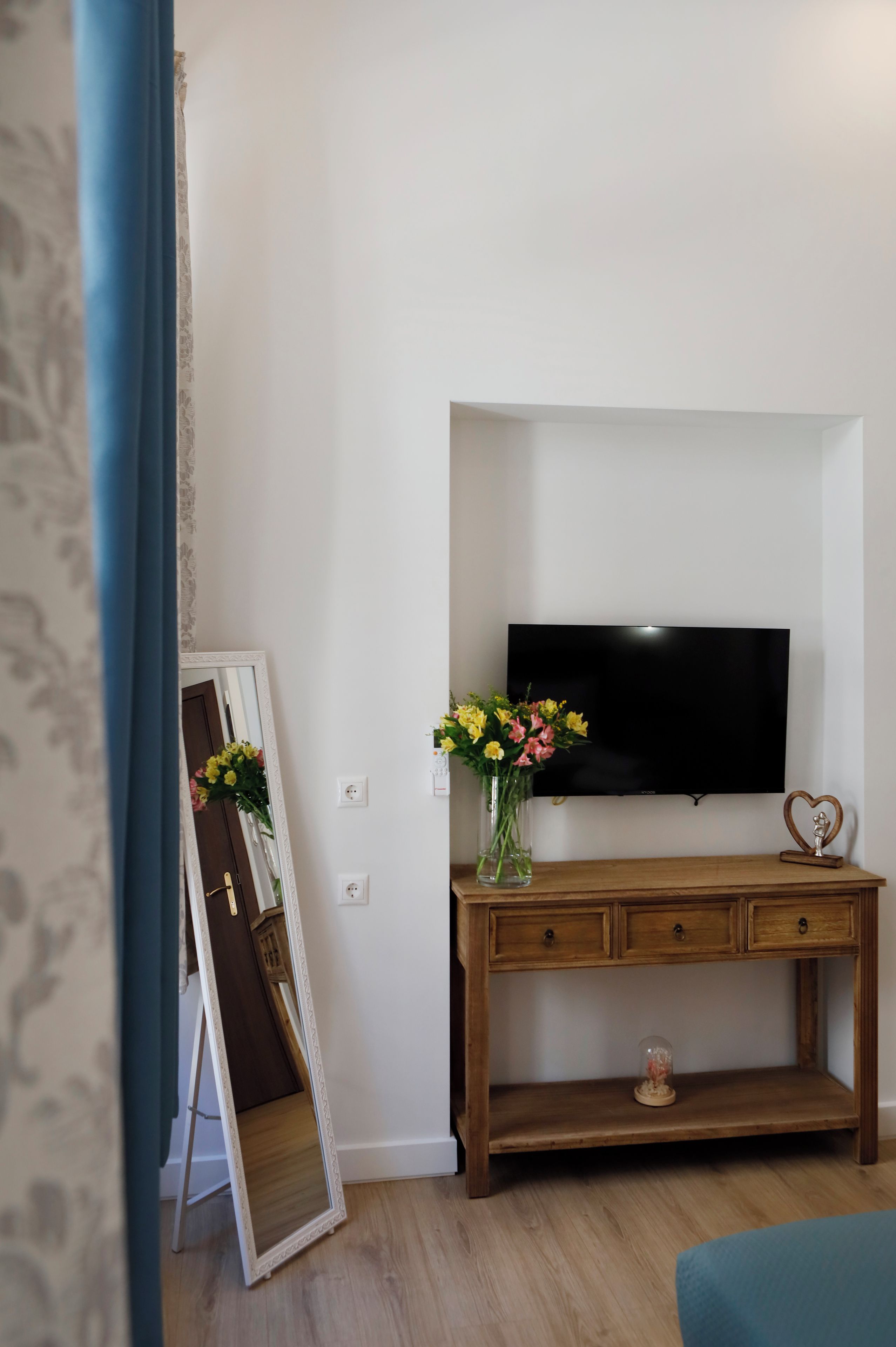 Una habitación con una consola de madera, un televisor de pantalla plana montado en la pared, un espejo apoyado contra la pared y un florero con flores sobre la mesa.