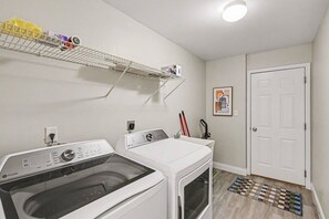 Lavandería con lavadora y secadora de carga frontal, un estante con productos de limpieza y una puerta con un cuadro enmarcado.