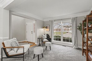 A living room with a sofa, armchair, ottoman, and a side table with a lamp. There is a large window with curtains, a door, and a bookshelf. - Freshly Renovated 4 Bedroom Home In Centerville OH (Dayton)
