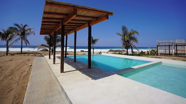 A beachside pool with a wooden pergola, palm trees, and a clear view of the ocean. - Pacific Dreams Hotel Suites  (Agua Blanca, Cozoaltepec)