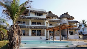 A multi-story building with a pool, thatched roofs, and a palm tree. - Pacific Dreams Hotel Suites  (Agua Blanca, Cozoaltepec)