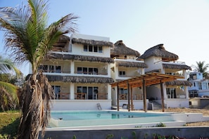 A multi-story building with a pool, thatched roofs, and a palm tree. - Pacific Dreams Hotel Suites  (Agua Blanca, Cozoaltepec)