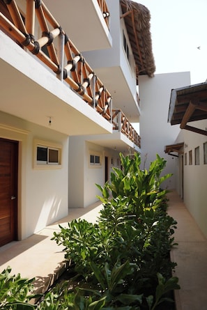 A building with a white facade, a wooden balcony with a thatched roof, and a narrow pathway lined with green plants. - Pacific Dreams Hotel Suites  (Agua Blanca, Cozoaltepec)