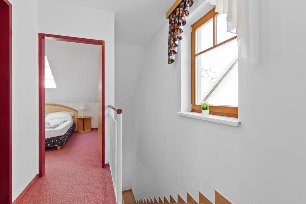 A corridor leading to a bedroom with a bed, a nightstand, and a window with a plant.