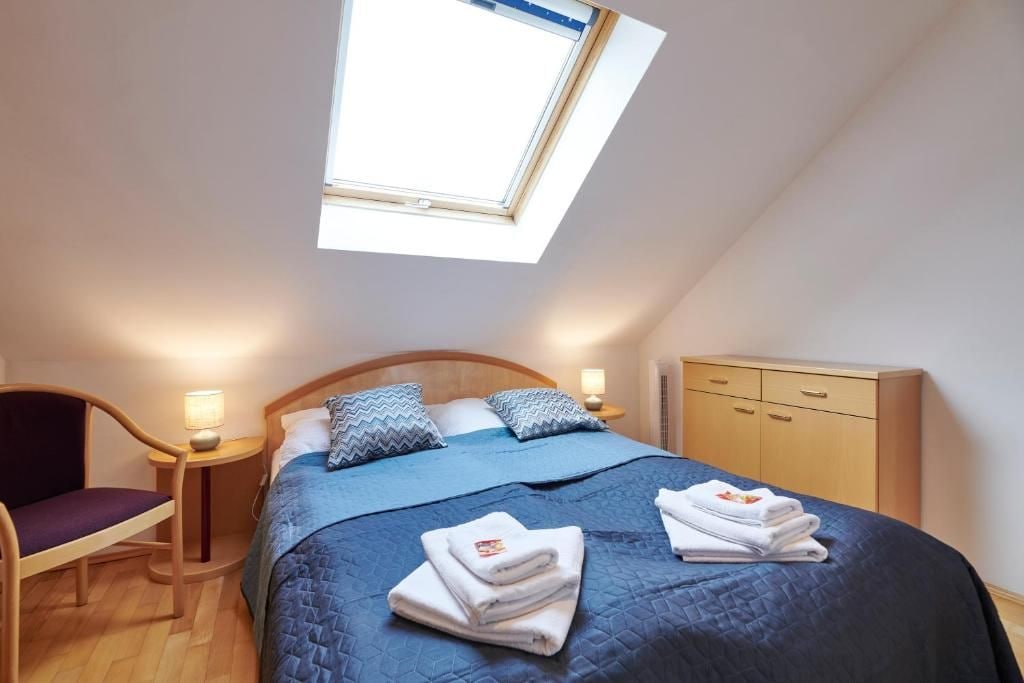Une chambre avec un puits de lumière, un lit avec une literie bleue, une table de chevet en bois avec une lampe, une armoire en bois et une chaise avec un coussin.