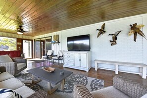 A spacious living room with a wooden ceiling, a flat-screen TV mounted on a white brick wall, wicker furniture, and a bench. - Point of View l 3BR - Dock & Big Views (Northport)