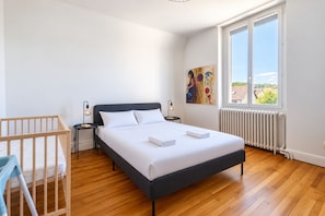 A modern bedroom with a large bed, a wooden crib, a nightstand, a wall-mounted lamp, and a window with a view of the outside. - The House of Burgundy in downtown Dijon (Dijon)