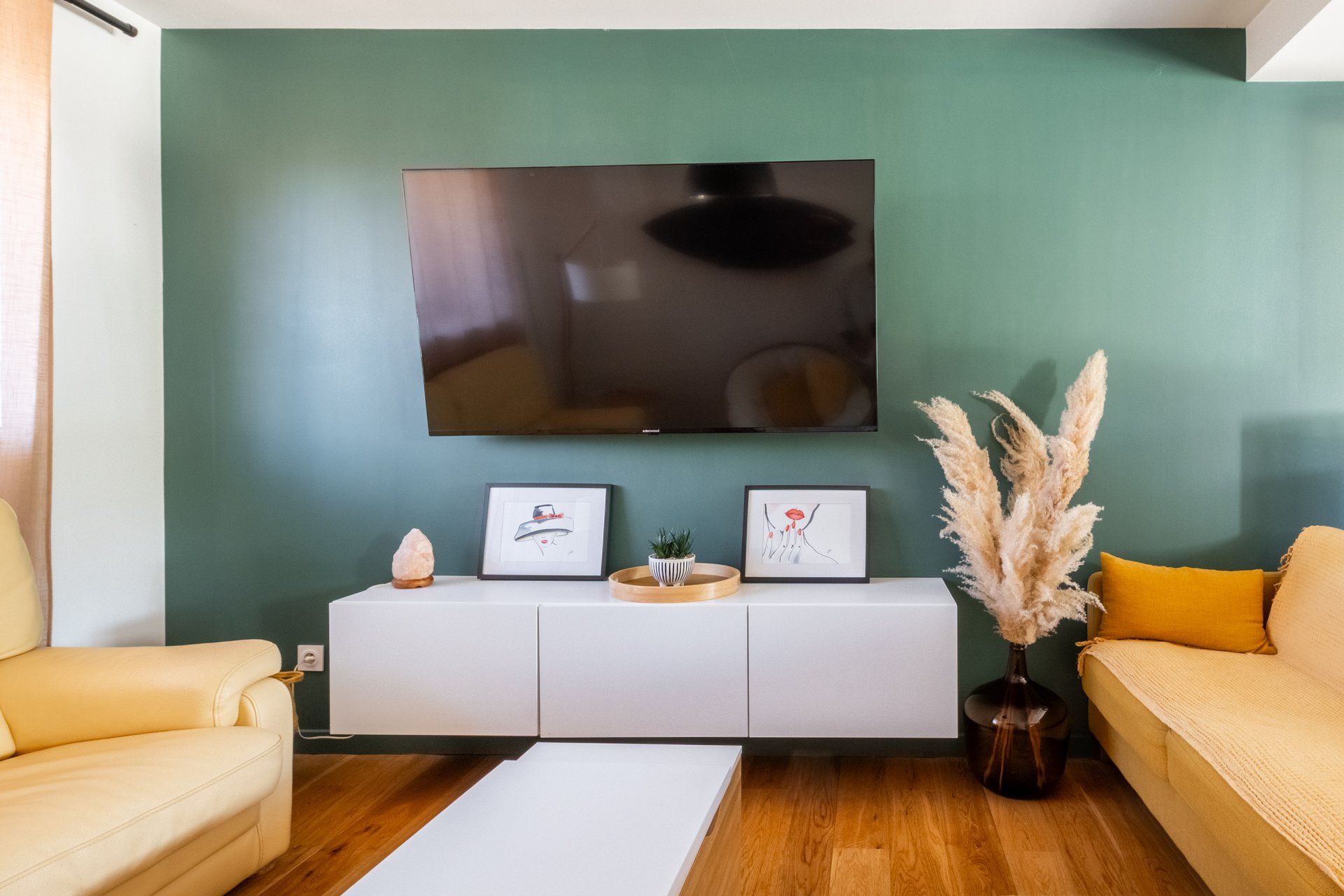 A living room with a flat-screen TV mounted on the wall, a white cabinet with decorative items, a sofa, and a wooden floor.