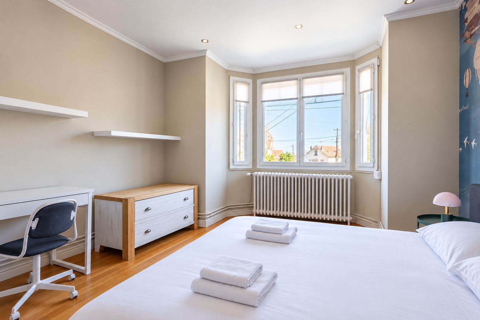 Ein Schlafzimmer mit Bett, Schreibtisch, Stuhl, Heizkörper und einem Fenster mit Jalousien.